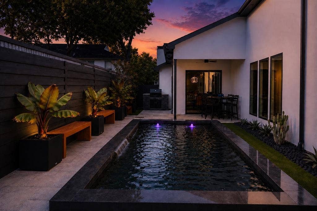 939 N Buckner Boulevard, Dallas, TX 75218 Private backyard featuring a rectangular pool with water features, a covered patio, an outdoor kitchen, a dark-stained privacy fence, and a landscaped perimeter