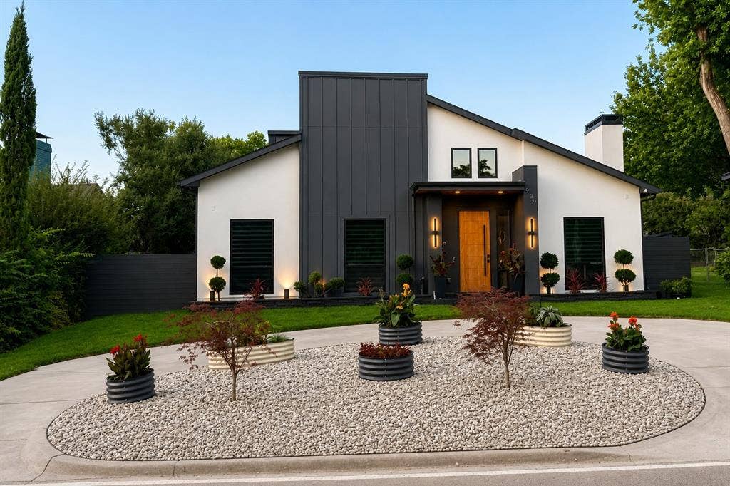 939 N Buckner Boulevard, Dallas, TX 75218 Modern exterior featuring a white stucco finish with dark vertical siding accents, a natural wood-tone entry door, and an aggregate circular driveway with a gravel landscape island