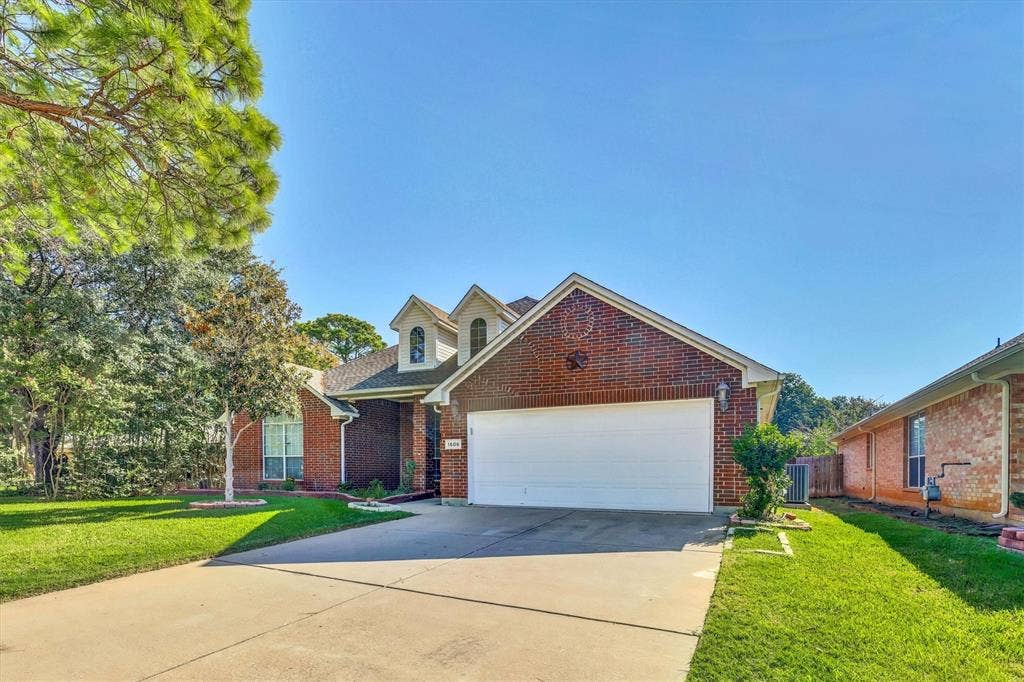 1506 Park Grove Drive, Irving, TX 75060 Brick exterior with a two-car garage, mature trees, a well-maintained lawn, and a concrete driveway