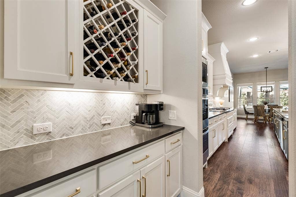 Built-in wine storage, white cabinetry with gold-tone hardware, dark countertops, herringbone tile backsplash, and recessed lighting