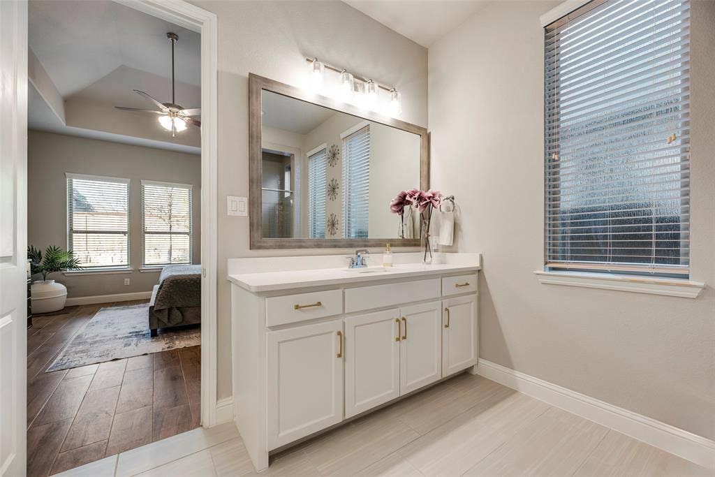 Vanity with a white countertop and gold-toned hardware, large framed mirror, and a window with blinds