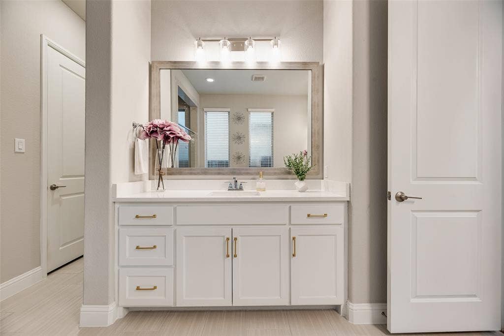 Single vanity with white cabinetry and gold-finish hardware, rectangular undermount sink, and light-tone countertop