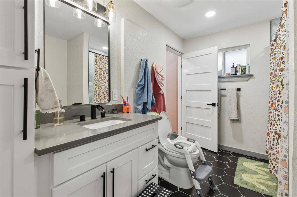 2408 Grandview Drive, Plano, TX 75075 White vanity with a light gray countertop and black fixtures, framed mirror with overhead vanity lighting, dark hexagonal floor tiling, and a white interior door with black hardware