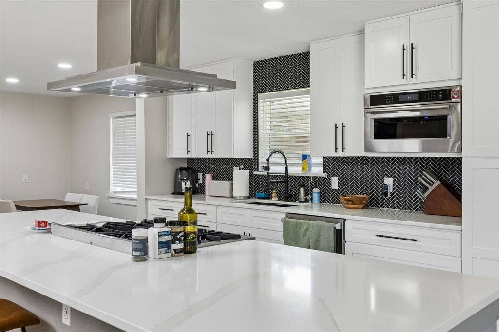 2408 Grandview Drive, Plano, TX 75075 Kitchen featuring a central island with a built-in cooktop, stainless steel range hood, white shaker cabinetry with dark hardware, and a black herringbone tile backsplash