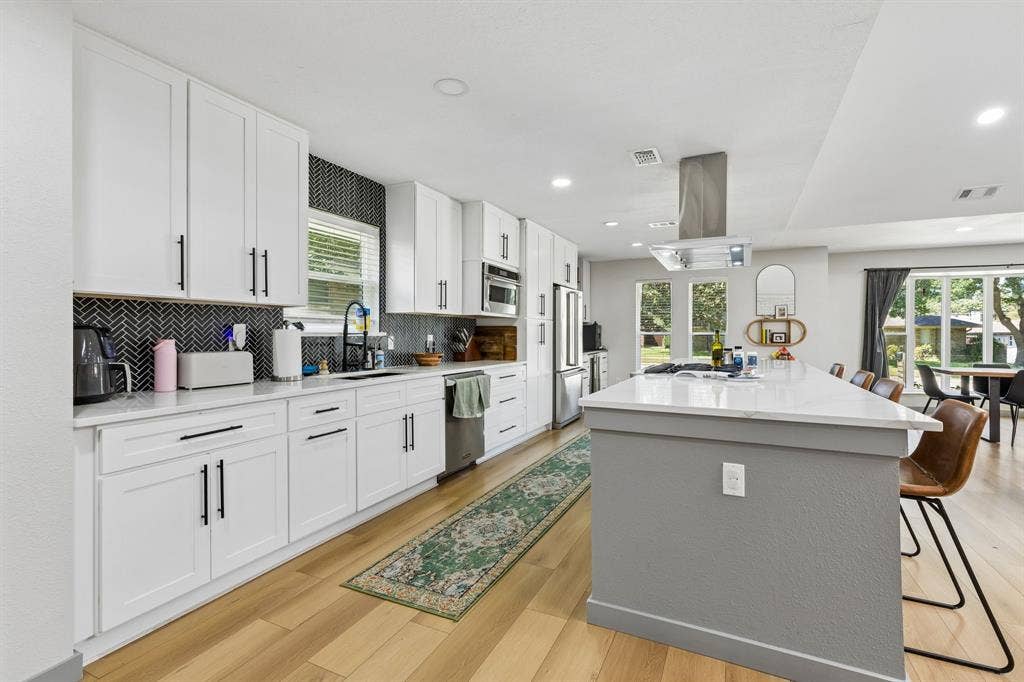 2408 Grandview Drive, Plano, TX 75075 Contemporary kitchen featuring white cabinetry, a black herringbone tile backsplash, an expansive island with a light-toned countertop, and wood-finish flooring