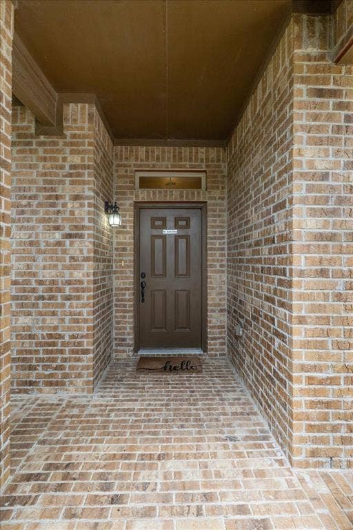 1713 Falling Star Drive, Fort Worth, TX 76052 Entryway featuring brick-clad walls and flooring, a brown paneled door with a transom window, and an attached wall sconce