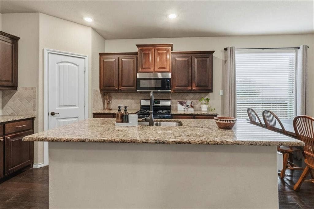 1713 Falling Star Drive, Fort Worth, TX 76052 Kitchen island with a built-in sink, granite countertops, dark wood-finish cabinetry, stainless steel appliances, and tile backsplash