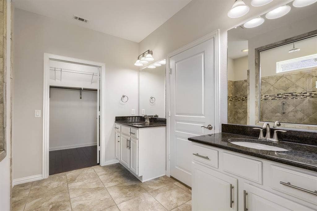 Dual vanity with white cabinetry and dark countertops, large mirror, recessed shower with tiled walls and glass enclosure, and a walk-in closet with built-in shelving