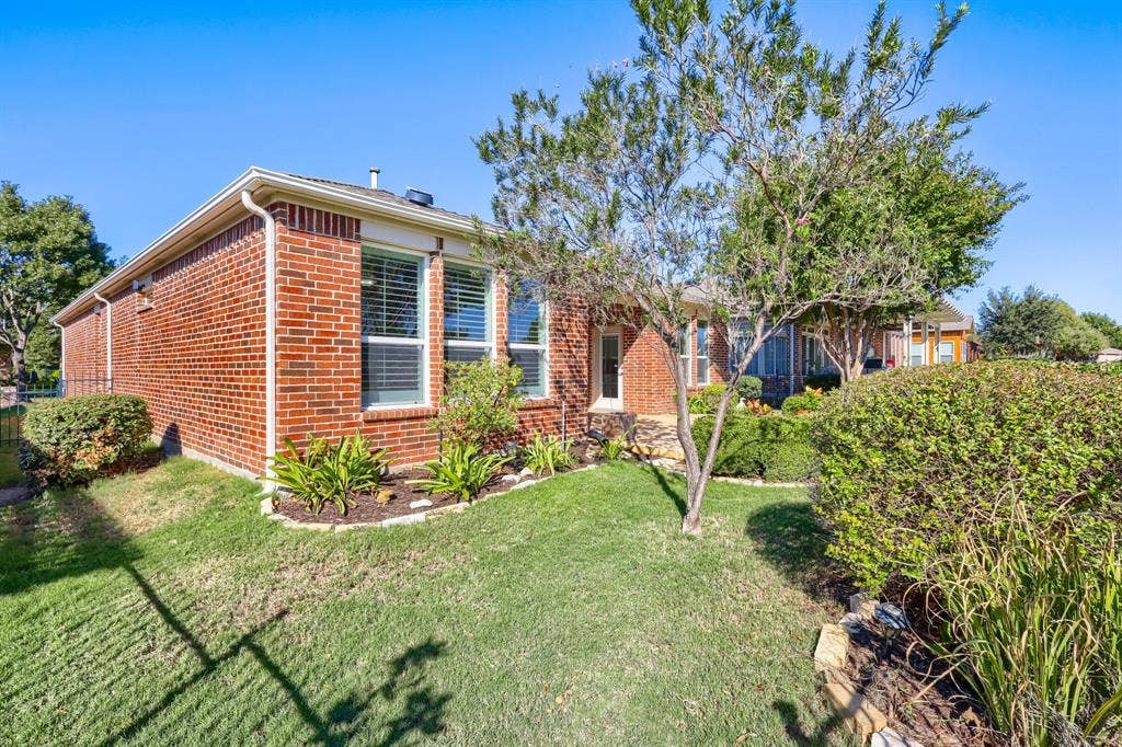 Red brick exterior with white window trim, multiple windows with horizontal blinds, well-maintained lawn, established trees, and perimeter landscaping