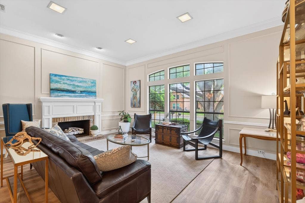 Living area featuring wood-finish flooring, a white mantel fireplace with brick surround, large windows, recessed lighting, and wall panel molding