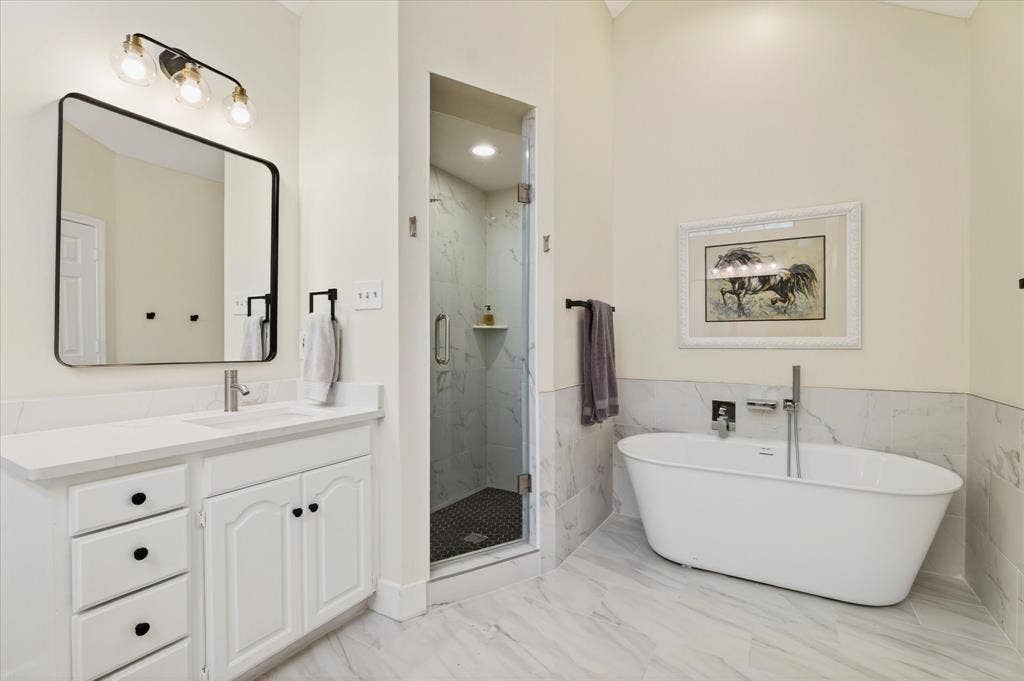 White vanity with a solid surface countertop, separate glass-enclosed shower with tiled walls, freestanding soaking tub, and large format floor tiling