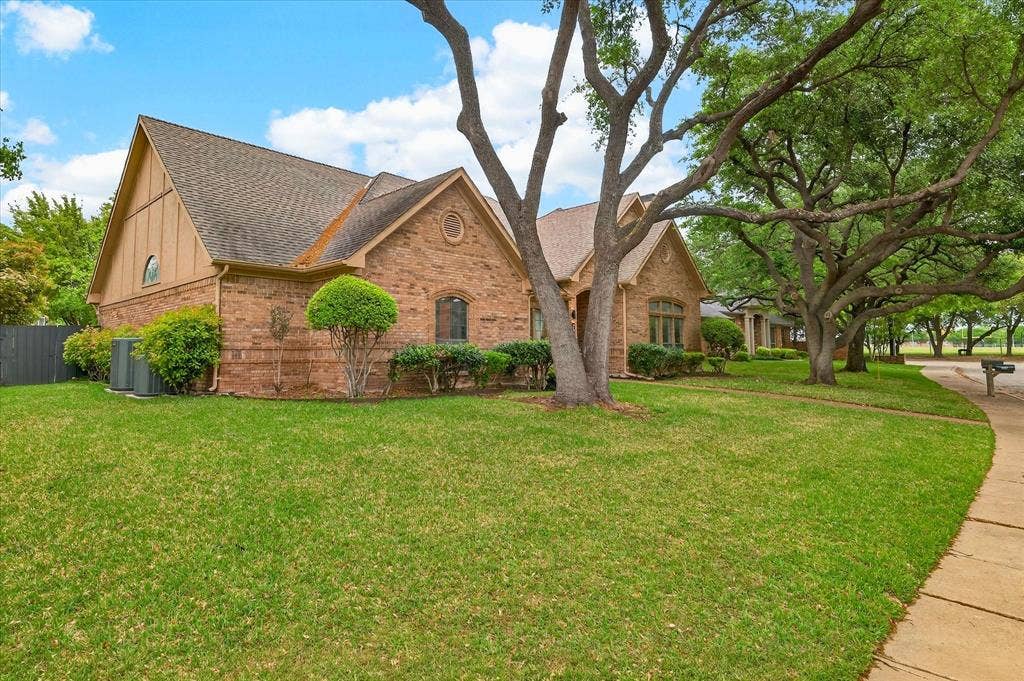 Brick exterior with gabled roof lines, mature trees, landscaped shrubbery, and a concrete sidewalk