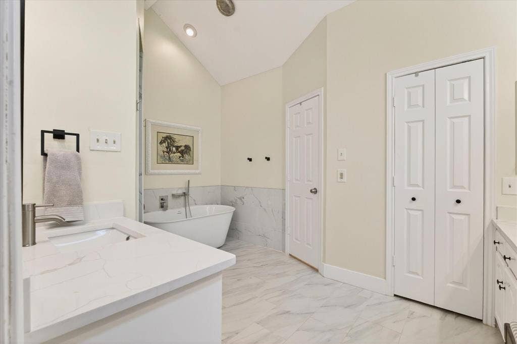 Freestanding soaking tub with wall-mounted faucet, marble-look tiled floors, white vanity with integrated sink and stone-look countertop, and recessed lighting