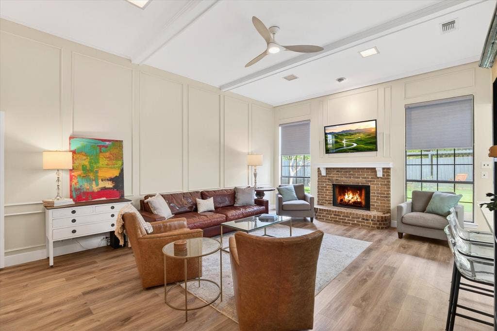 Vaulted ceiling with a contemporary ceiling fan, wood-finish flooring, brick fireplace with a white mantel, paneled accent wall, and multiple windows