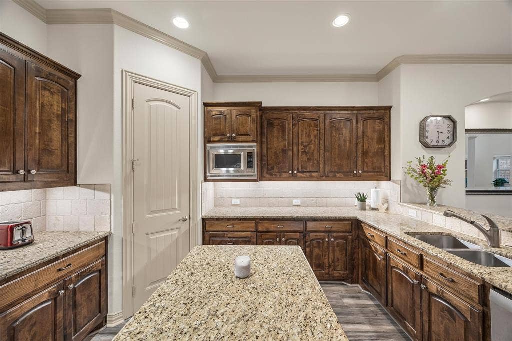 8249 Short Street, Frisco, TX 75034 Kitchen featuring wood-finish cabinetry, light speckled countertops, a double basin stainless steel sink, and a tiled backsplash