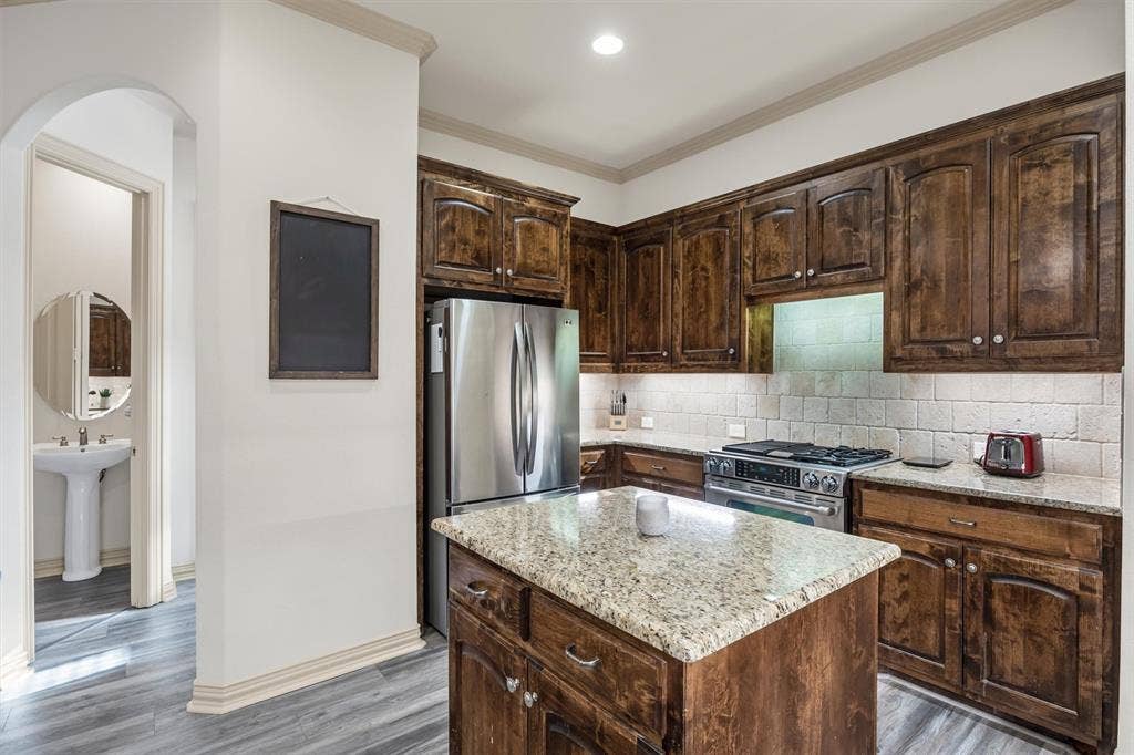 8249 Short Street, Frisco, TX 75034 Kitchen featuring wood-finish cabinetry, stainless steel appliances, a stone countertop island, stone countertops, and a subway tile backsplash