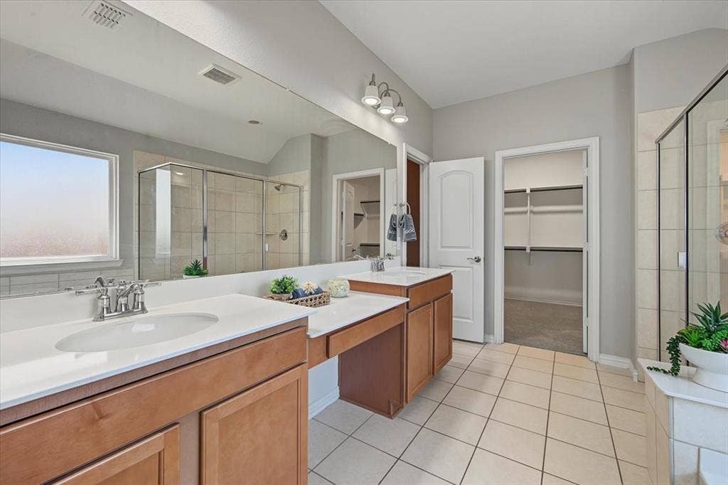 Dual vanities with white countertops, a large framed mirror, tiled flooring, and a glass-enclosed shower