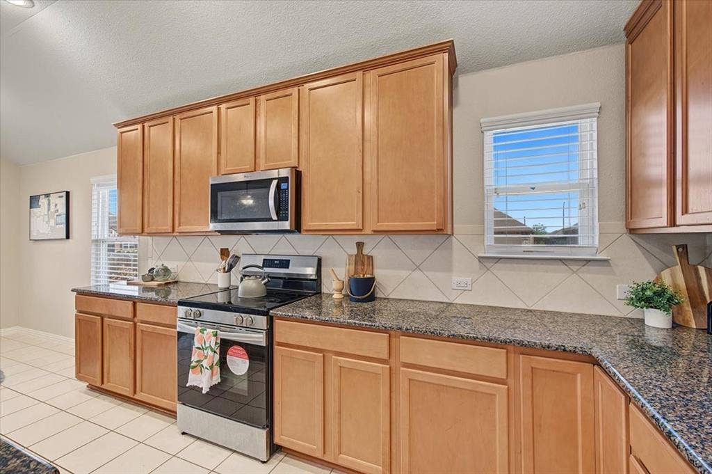 Kitchen featuring wood-finish cabinetry, speckled countertops, diagonal tile backsplash, stainless steel appliances, and tile flooring