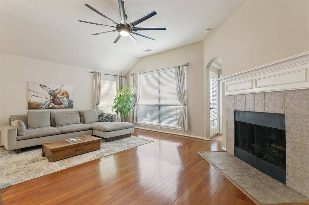 Varnished wood-finish flooring, vaulted ceiling with a modern ceiling fan, large windows with blinds, tiled fireplace with a white mantel, and an arched doorway