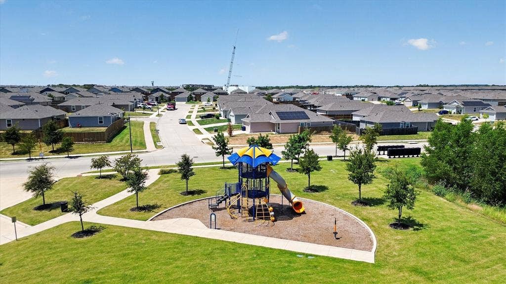 Community green space featuring a playground with slides and climbing elements, surrounded by a manicured lawn, paved walkways, and new construction homes