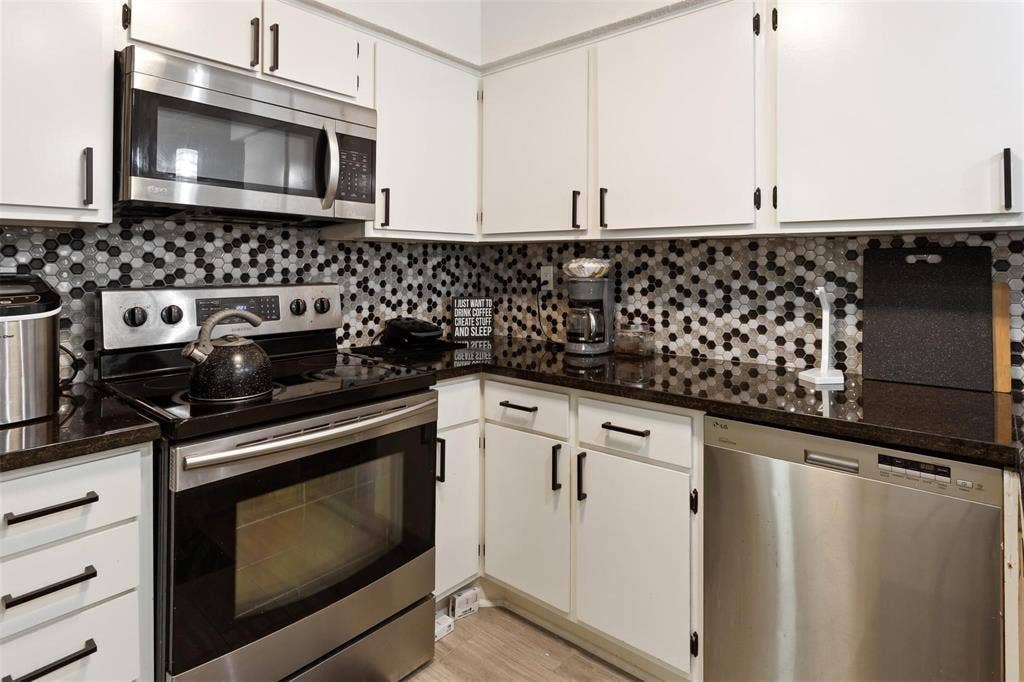 13026 Leader Street, #951, Houston, TX 77072 White cabinetry with matte black hardware, dark countertops, hexagonal mosaic tile backsplash, stainless steel appliances, and wood-finish flooring