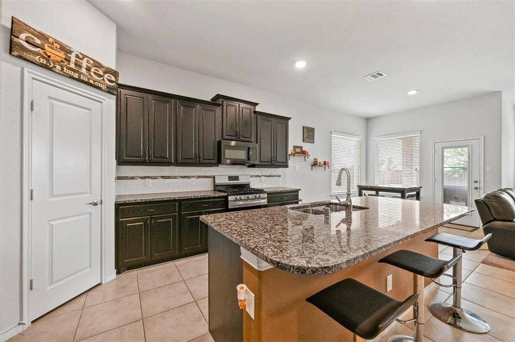 602 Brock Drive, Anna, TX 75409 Kitchen featuring dark wood-finish cabinetry, stainless steel appliances, a multi-tone tile backsplash, and an island with a polished stone countertop and integrated sink