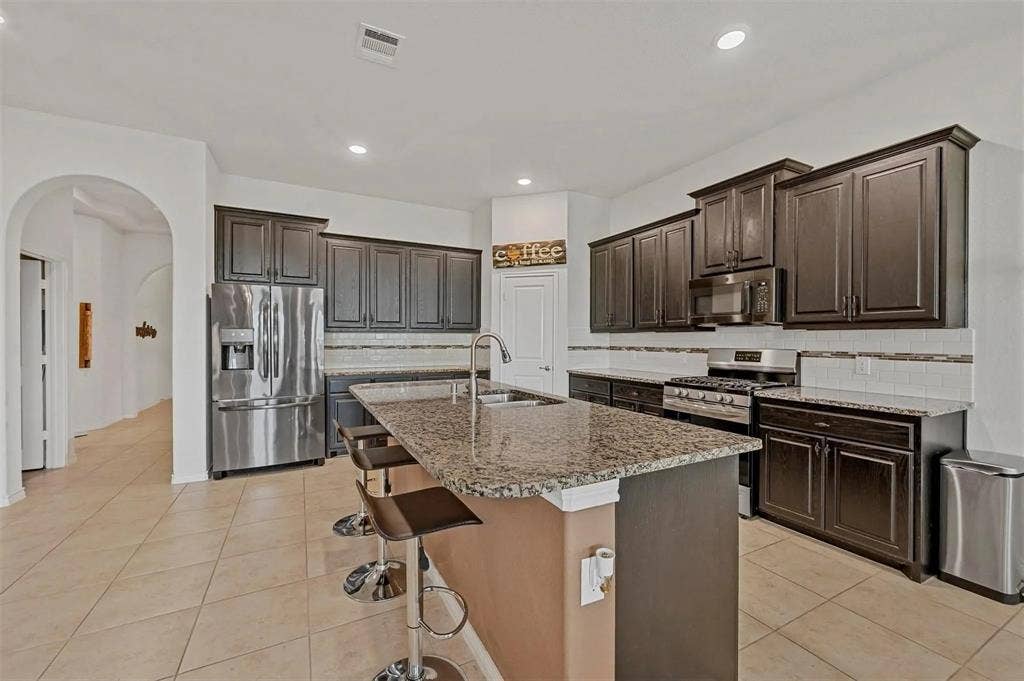 602 Brock Drive, Anna, TX 75409 Kitchen featuring dark wood-finish cabinetry, stainless steel appliances, granite countertops, a large kitchen island with a sink, and tile flooring