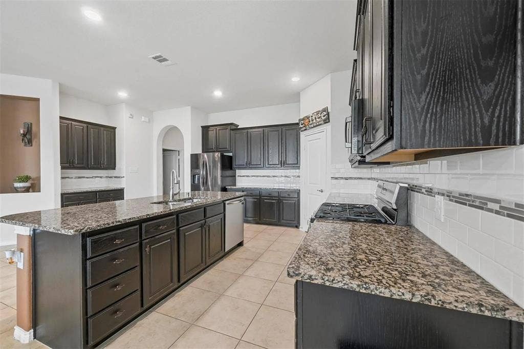 602 Brock Drive, Anna, TX 75409 Kitchen featuring dark wood-finish cabinetry, granite countertops, tile flooring, stainless steel appliances, and an expansive center island with an undermount sink