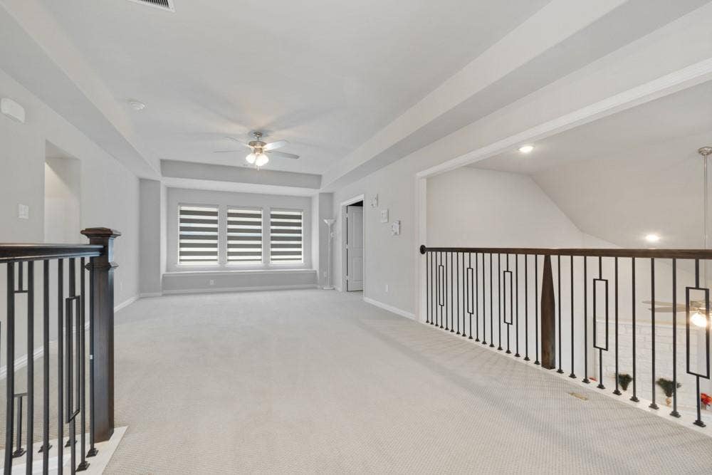 Spacious carpeted landing featuring dark wood newel posts, wrought iron balusters, a ceiling fan with integrated lighting, and a window with horizontal blinds