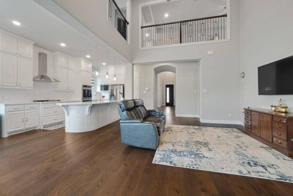 Open-concept living area featuring wood-finish flooring, a kitchen with white cabinetry and an island, a stainless steel range hood, and a second-level open railing