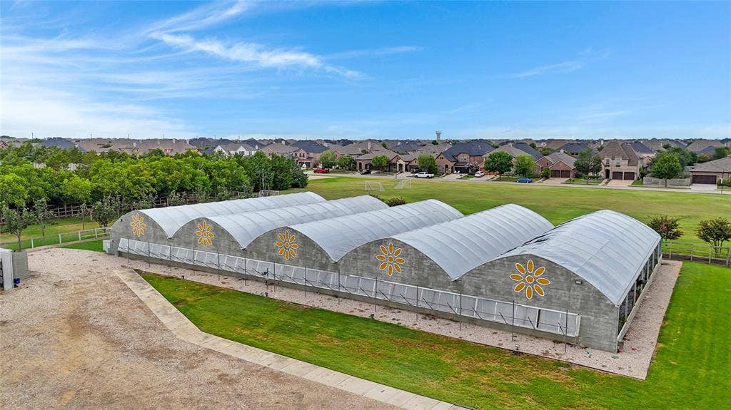 Multiple arched greenhouse structures with decorative flower motifs, surrounded by a gravel perimeter, an expansive grass lawn, and mature trees