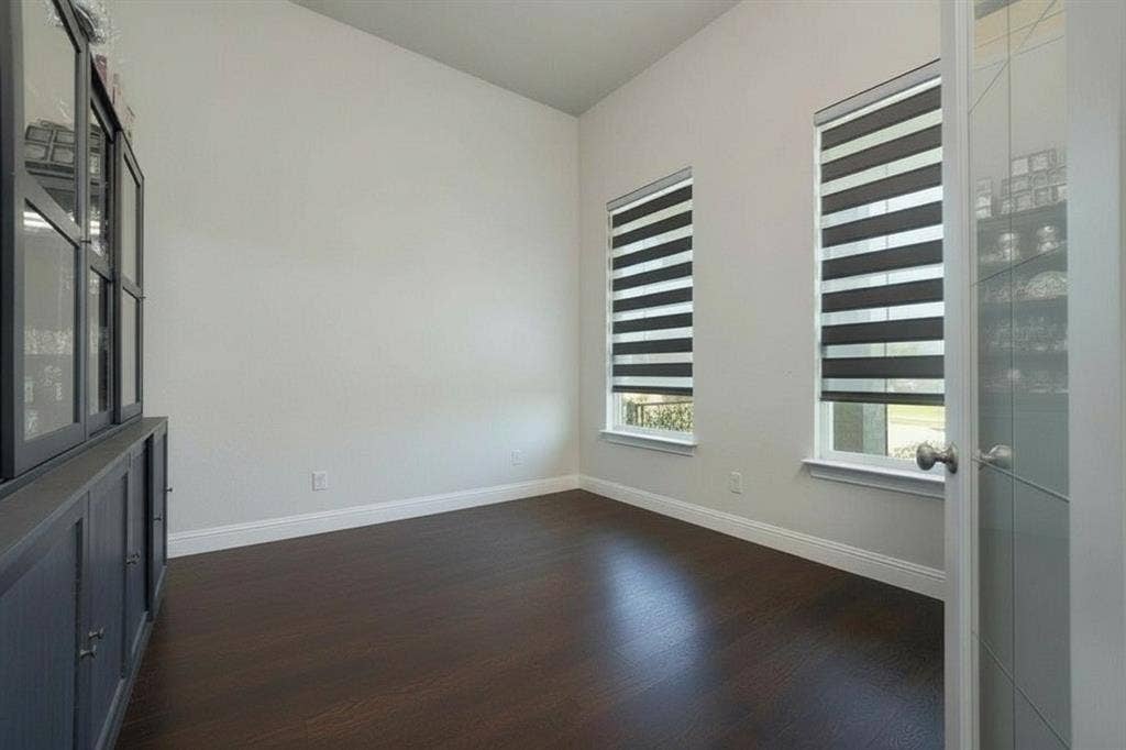 Vaulted ceiling, dark wood-finish flooring, two windows with striped roller shades, built-in cabinetry with glass-front uppers, and a paneled glass door