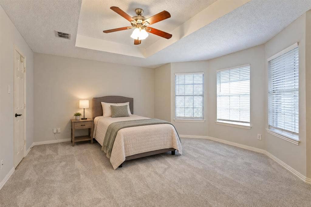 Tray ceiling with a central multi-light ceiling fan, neutral wall tones, carpeted flooring, and a bay window ( virtually Staged)