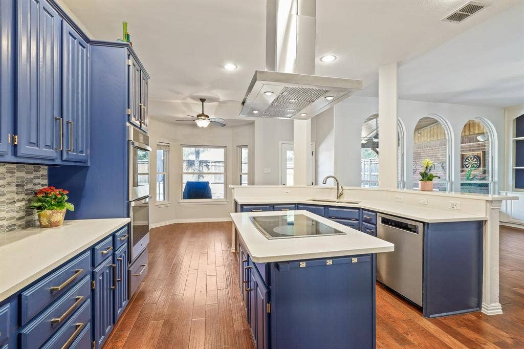 Kitchen featuring blue cabinetry, a central island with a built-in cooktop, stainless steel range hood, wood-finish flooring, and arched windows