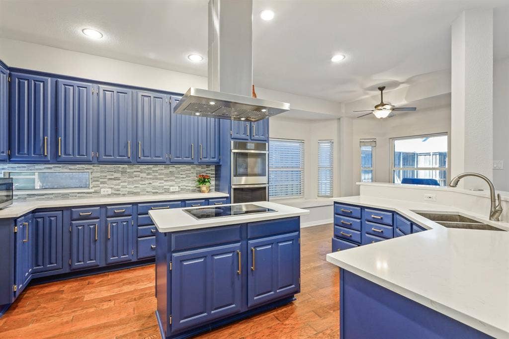 Kitchen featuring custom blue cabinetry, light-toned solid surface countertops, a stainless steel chimney-style range hood, integrated wall oven, and wood-finish flooring