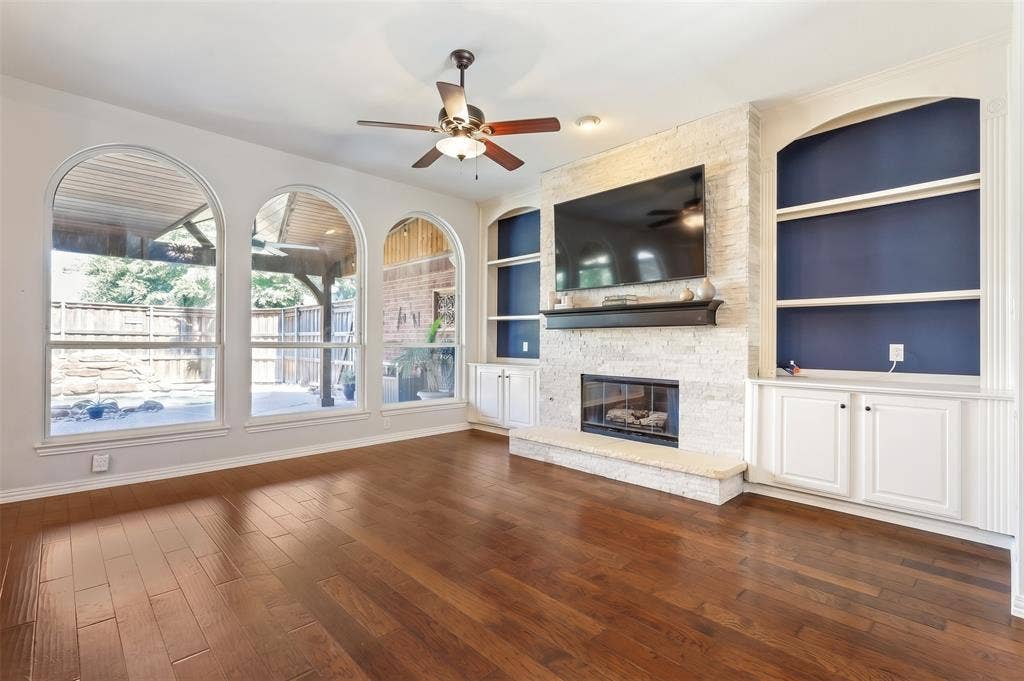 Living area featuring wood-finish flooring, floor-to-ceiling stone fireplace, built-in cabinetry with arched shelving, and triple arched windows