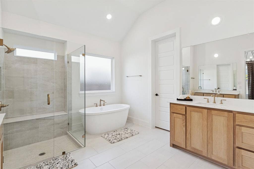 1117 Bradford Street, Celina, TX 75009 Bathroom featuring a freestanding soaking tub, glass-enclosed shower with bench seating, dual vanity with wood-finish cabinetry and light-toned countertops, and light-toned tile flooring