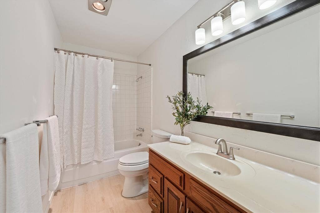 Bathroom featuring a single-sink vanity with wood-finish cabinetry and new lighting, an integrated basin, a large framed mirror, light-tone wood-finish LVP flooring, and a tub-shower combination with tiled surround