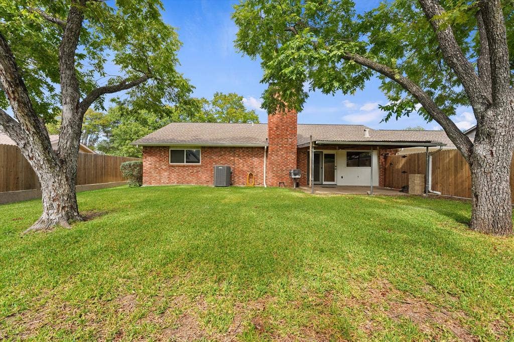 Exterior featuring brick siding, a covered patio, a brick chimney, and mature trees situated on a lawn enclosed by wood fencing