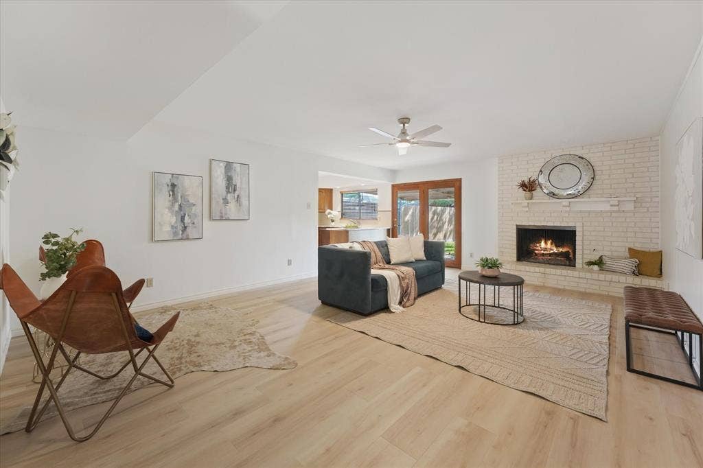 Expansive living area ready to entertain, featuring wood-finish LVP flooring, a white brick fireplace with a built-in hearth, a ceiling fan, and glass-paneled wooden exterior doors