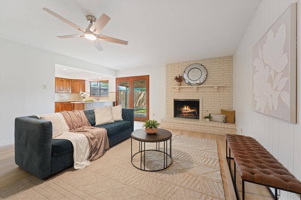 Living area featuring a floor-to-ceiling brick fireplace with a raised hearth, wood-finish LVP flooring, a multi-blade ceiling fan with integrated lighting, and glass-paneled patio doors.  Ready for a cool fall evening in front of the fireplace.