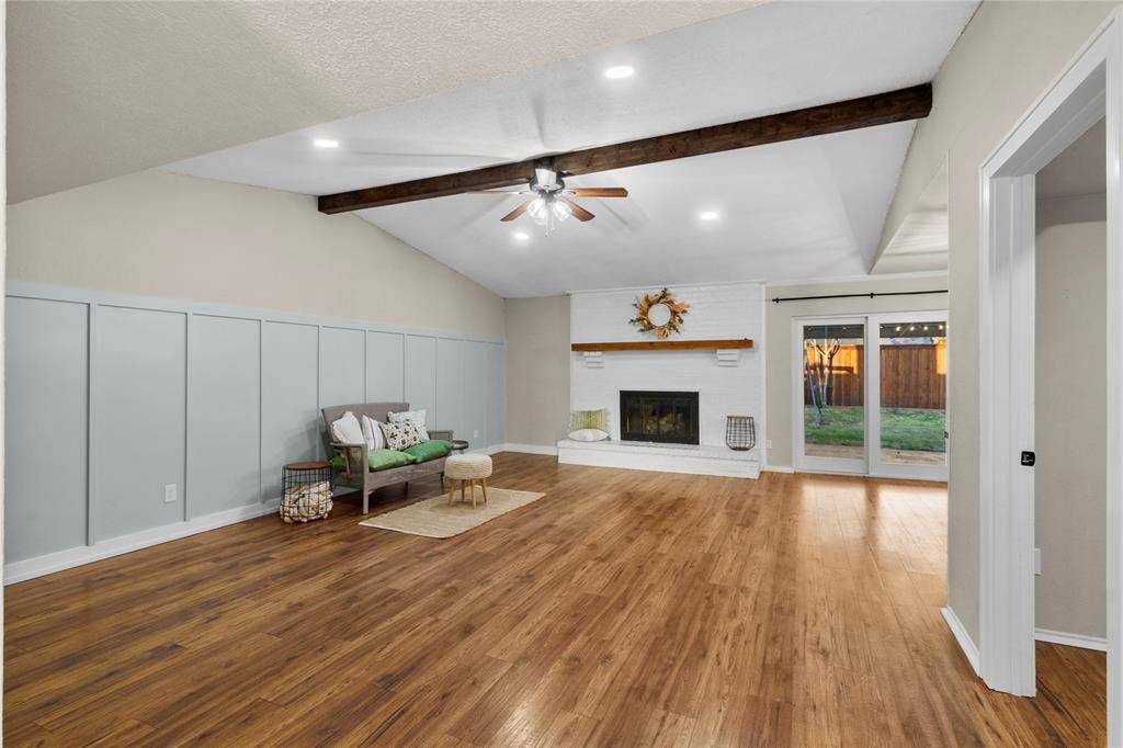 Vaulted ceiling with exposed beam, recessed lighting, wood-finish flooring, white brick fireplace with wood mantel, and board and batten wall accent
