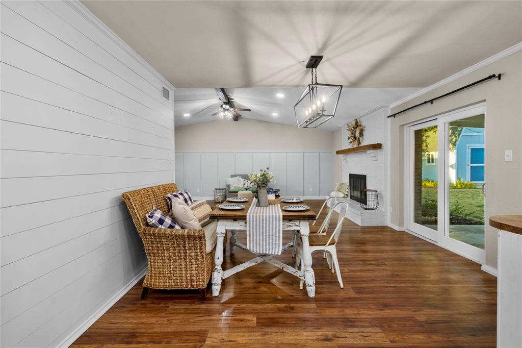 Warm wood-finish flooring, white shiplap accent wall, modern linear chandelier, white brick fireplace with a wood mantel, and sliding glass patio doors
