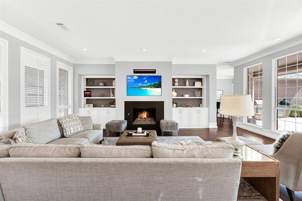 5325 BLAKE Drive, Plano, TX 75093 A sleek black fireplace hearth flanked by custom inset shelving units forms the visual anchor of this bright family space. Wall-to-wall windows paired with white plantation shutters flood the room with natural light while offering views of the covered patio.