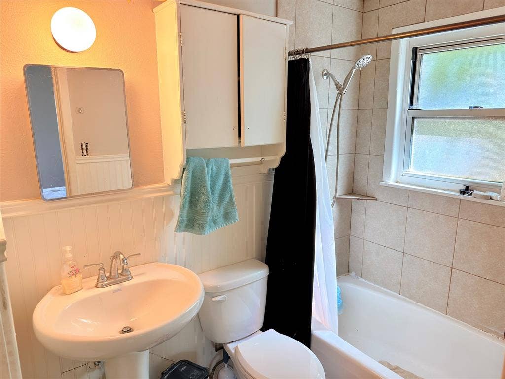 3014 Crestview Street, Tyler, TX 75701 Pedestal sink with chrome fixtures, wall-mounted medicine cabinet, beadboard wainscoting, tiled bath enclosure with built-in shelving, and a window for natural light