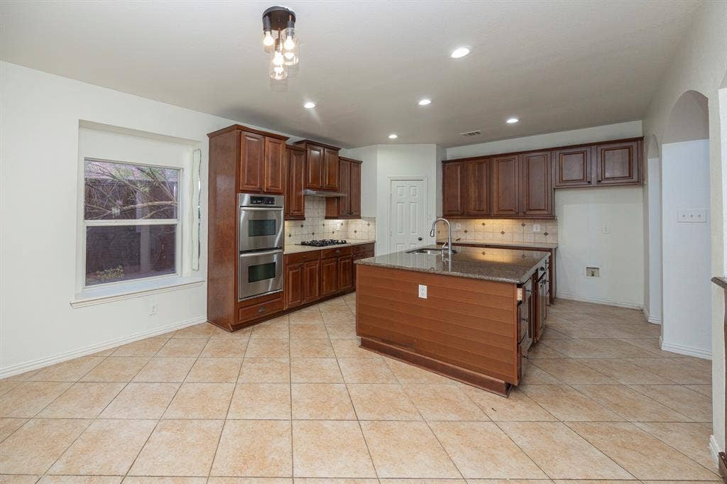 6405 Sapphire Drive, McKinney, TX 75070 Spacious kitchen featuring an island with a built-in sink, stainless steel double ovens, rich wood-finish cabinetry, tile flooring, and recessed lighting