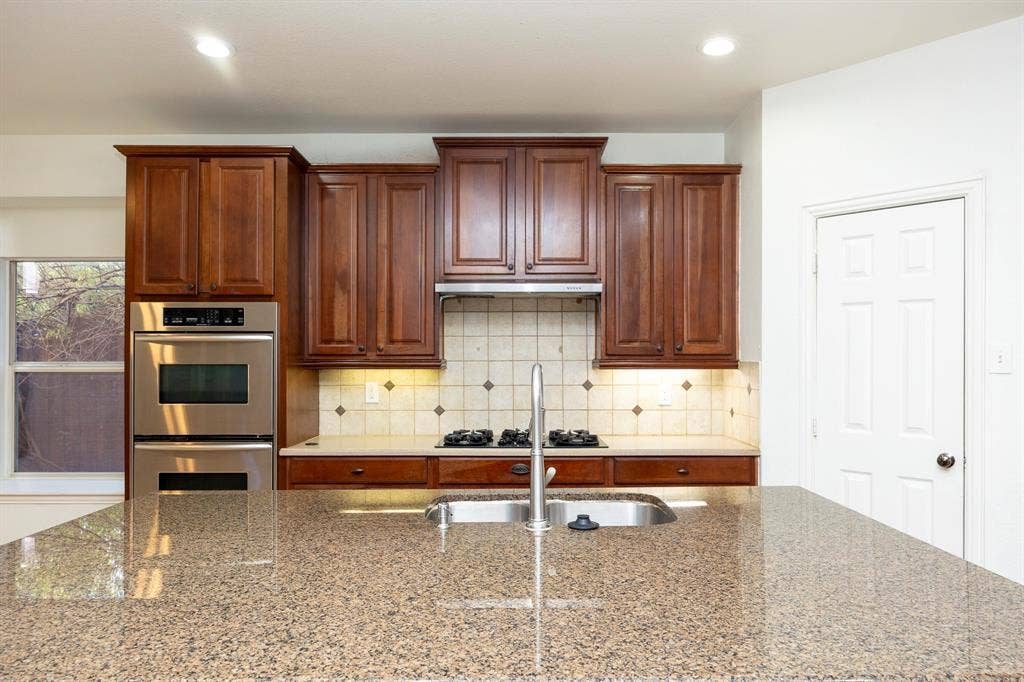 6405 Sapphire Drive, McKinney, TX 75070 Kitchen featuring an expansive stone countertop, stainless steel double oven, gas cooktop, tile backsplash, and wood cabinetry