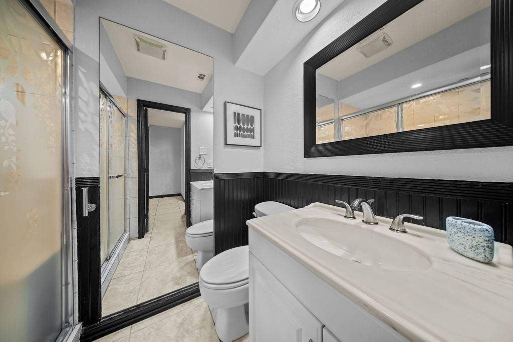 9302 Chimney Sweep Lane, Dallas, TX 75243 White vanity with an integrated sink and brushed nickel faucet, large framed mirror, vertical wall paneling, and a shower with frosted glass doors