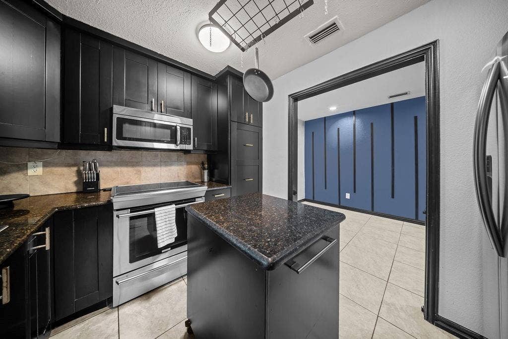 9302 Chimney Sweep Lane, Dallas, TX 75243 Black cabinetry paired with granite countertops, stainless steel appliances, a tiled backsplash, and light-toned floor tiles