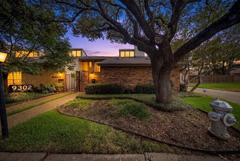 9302 Chimney Sweep Lane, Dallas, TX 75243 Brick exterior with upper-level windows, mature trees, landscaped front yard, and a paved walkway leading to a gated entry